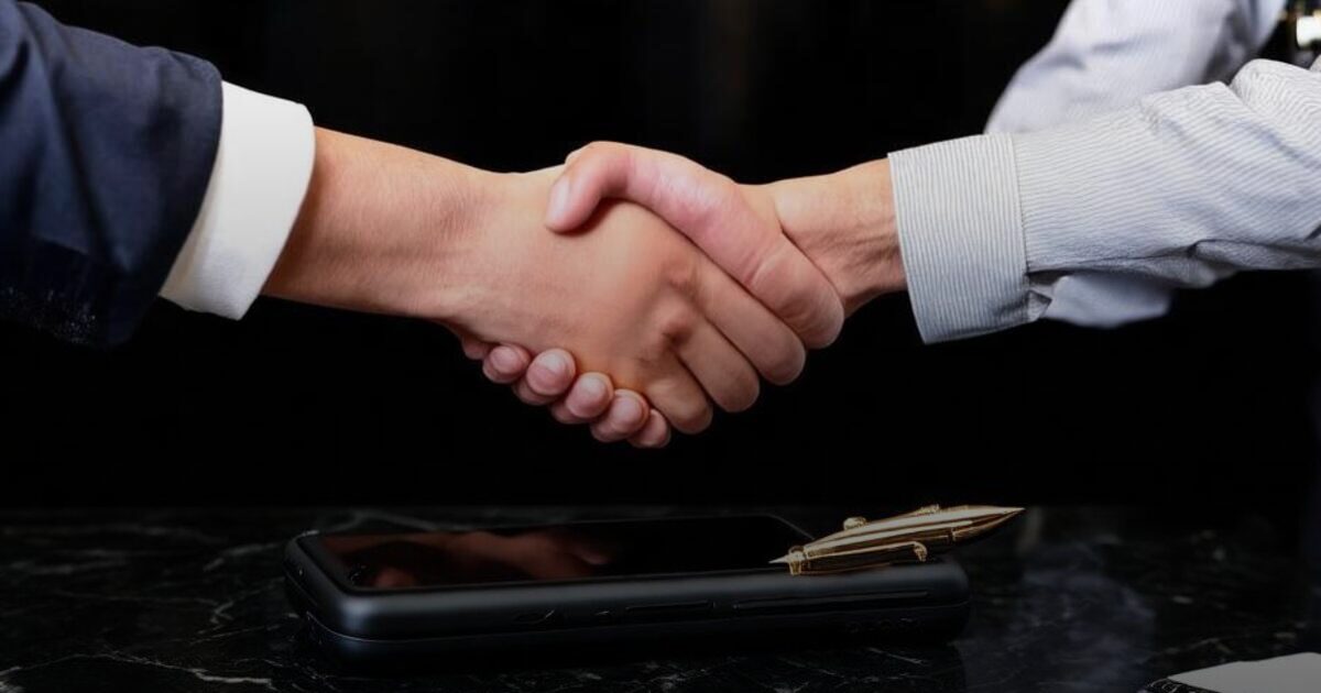 Two people shaking hands over a dark marble table beside a face-down phone and a gold pen
