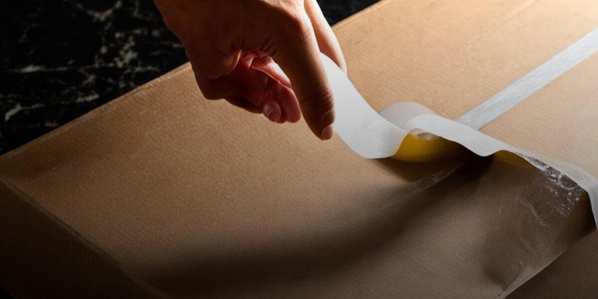 A hand smoothing down a strip of clear packing tape on a plain cardboard shipping box, part of expanding a listing onto a second marketplace