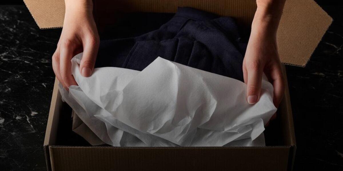 Hands folding tissue paper over a garment inside an open cardboard shipping box