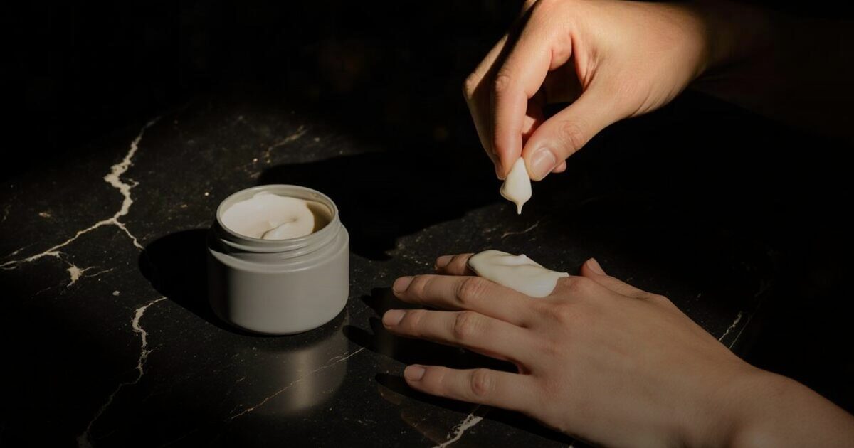 Two hands scooping cream from a plain ceramic jar and smoothing it onto skin